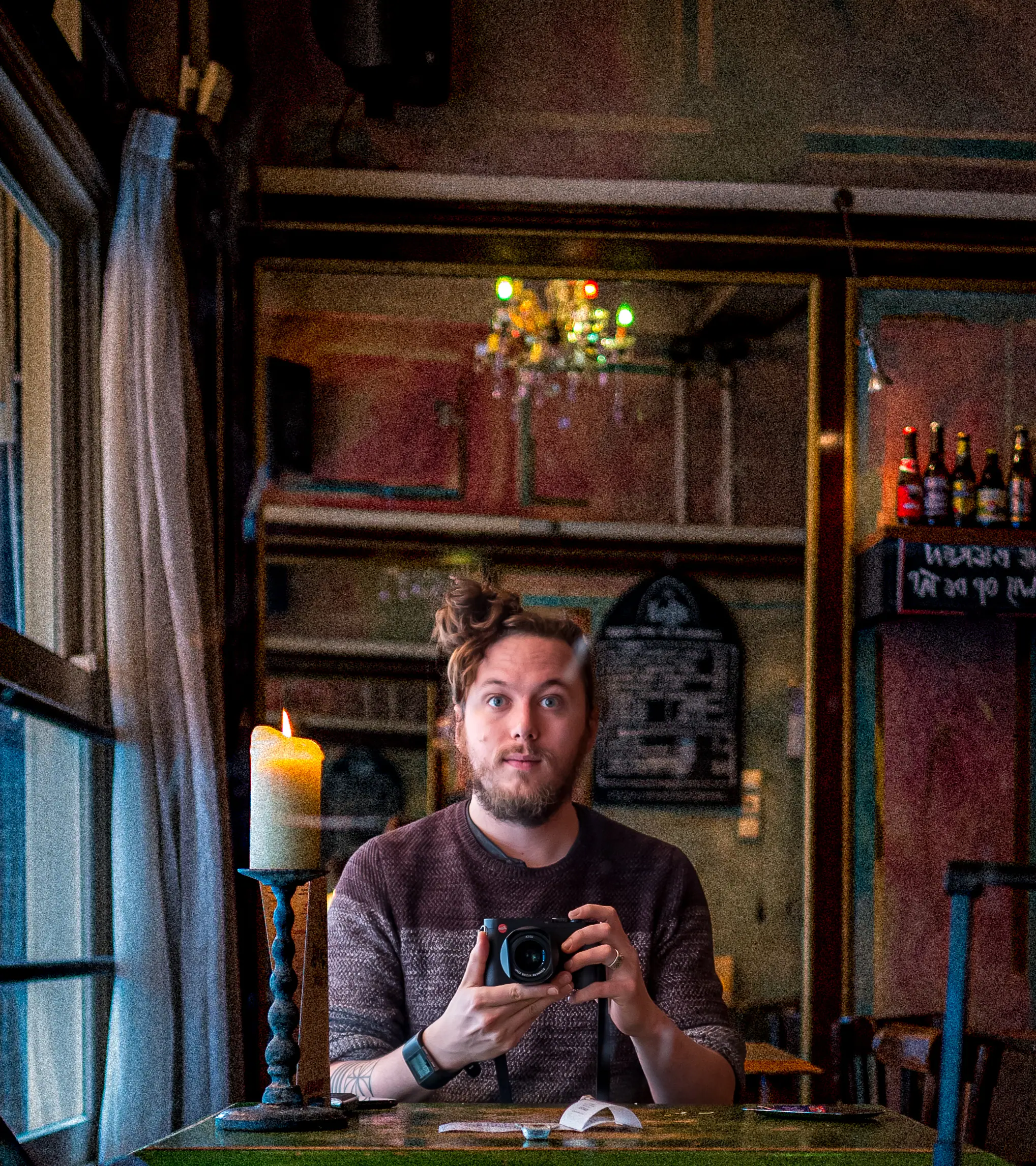 I'm facing a mirror while sitting at a table with a candle beside me. My Leica camera is pointing at the mirror, as I hold it at chest height. My face is neutral, my hair is up in a messy bun. A typical brown bar setting in the Netherlands with wooden surroundings, beer bottles with the special drinks listed in the top right corner, and on the left is an open window letting in the natural light.